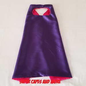 May include: A reversible cape with a deep purple exterior and a bright pink interior. The cape has a neck opening and is made of a shiny, satin-like material. The text "SUPER CAPES AND MORE" is printed at the bottom.