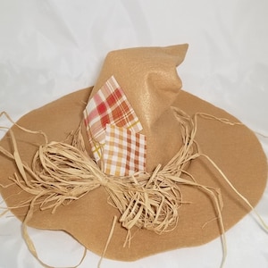 May include: A brown felt scarecrow hat with a wide brim and a pointed crown. The hat is decorated with plaid fabric patches in shades of red, orange, and white. The brim is trimmed with straw.