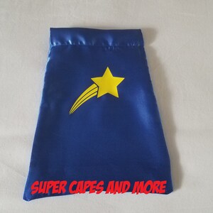 May include: Blue satin cape with a yellow star and a shooting star design. The cape has the text "SUPER CAPES AND MORE" printed on the bottom.