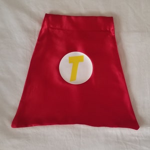 May include: Red satin cape with a white circle and a yellow letter T in the center.