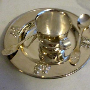 May include: A silver tea set including a small cup with a handle, a saucer, and two spoons. The set has a shiny finish and decorative floral accents around the saucer's edge. The cup is cylindrical and sits on the saucer.