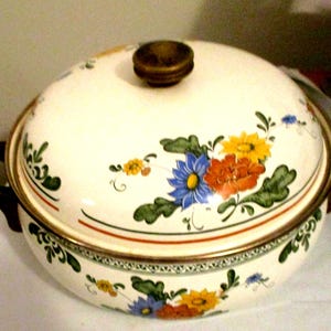 May include: Cream-colored enamelware casserole dish with lid. The dish features a floral design with blue, orange, and yellow flowers. It has brown handles and a brown knob on the lid. The rim has a gold trim.