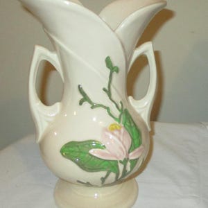 Peut inclure: Un vase en c&eacute;ramique vintage de couleur cr&egrave;me avec deux anses et un motif floral. Le vase pr&eacute;sente une fleur de magnolia rose avec des feuilles et des tiges vertes.