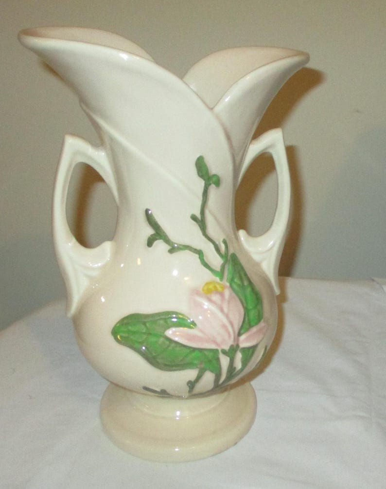 Peut inclure: Un vase en c&eacute;ramique vintage de couleur cr&egrave;me avec deux anses et un motif floral. Le vase pr&eacute;sente une fleur de magnolia rose avec des feuilles et des tiges vertes.