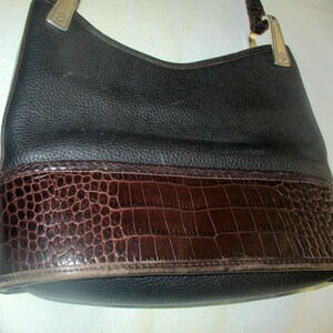 Puede incluir: Un bolso hobo de cuero negro con un acabado de grano texturizado en la parte superior y un panel de cuero marr&oacute;n oscuro con relieve de cocodrilo en la parte inferior. El bolso incluye herrajes met&aacute;licos para la correa y un asa trenzada.