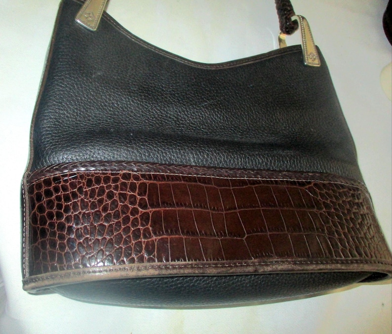 Puede incluir: Un bolso hobo de cuero negro con un acabado de grano texturizado en la parte superior y un panel de cuero marr&oacute;n oscuro con relieve de cocodrilo en la parte inferior. El bolso incluye herrajes met&aacute;licos para la correa y un asa trenzada.