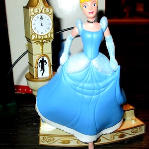 May include: A Cinderella figurine in a blue gown with a clock tower ornament. The clock tower is gold and cream-colored, with a clock face showing the time as a few minutes before midnight. The figurine is standing on a decorative base.