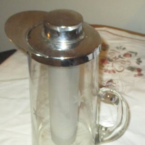 May include: A clear glass pitcher with a frosted glass insert and a silver lid. The pitcher has a handle and a decorative etched design.