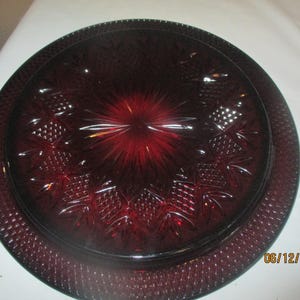 May include: A deep red, round glass serving plate with a textured rim and intricate pineapple and starburst patterns. The plate is approximately 12 inches in diameter, perfect for displaying food or as a decorative piece.