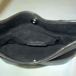 Puede incluir: El interior de un bolso de color marr&oacute;n oscuro con forro de tela negra, un bolsillo central con cremallera y un cierre de bot&oacute;n a presi&oacute;n plateado en el borde superior. El bolso tiene una abertura estructurada con costuras visibles.