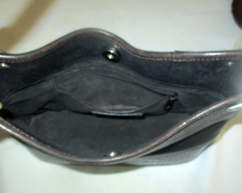 Puede incluir: El interior de un bolso de color marr&oacute;n oscuro con forro de tela negra, un bolsillo central con cremallera y un cierre de bot&oacute;n a presi&oacute;n plateado en el borde superior. El bolso tiene una abertura estructurada con costuras visibles.