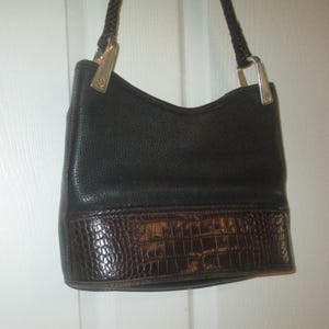 Puede incluir: Un bolso de mano de cuero negro con un acabado texturizado en la parte superior y un panel de cuero marr&oacute;n oscuro con relieve de cocodrilo en la base. El bolso incluye una correa de hombro de cuero trenzado con herrajes met&aacute;licos plateados.