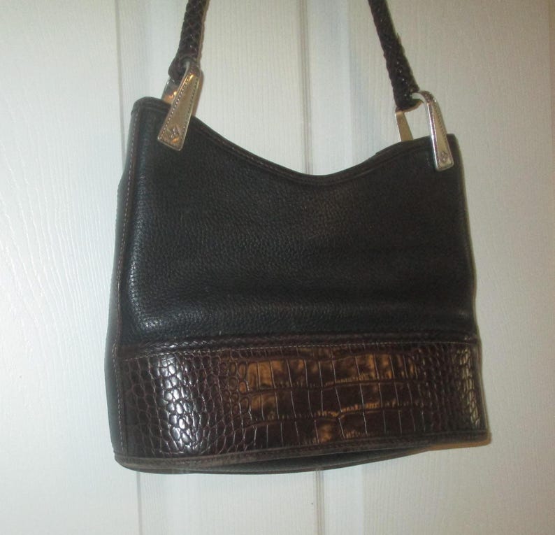 Puede incluir: Un bolso de mano de cuero negro con un acabado texturizado en la parte superior y un panel de cuero marr&oacute;n oscuro con relieve de cocodrilo en la base. El bolso incluye una correa de hombro de cuero trenzado con herrajes met&aacute;licos plateados.