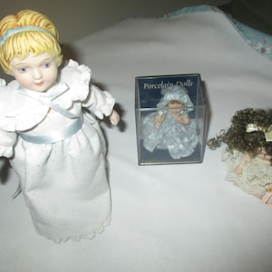 May include: A large porcelain doll with blonde hair and a blue headband wearing a white dress with a blue ribbon, a small doll inside a clear box labeled Porcelain Dolls, and a small doll with curly brown hair wearing a white lace dress.