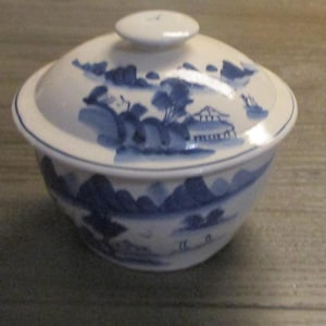 May include: A white ceramic bowl with a lid, featuring a blue landscape design. The bowl has a round shape with a lid and a small knob. The landscape design includes mountains, trees, and buildings.