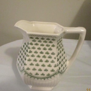 May include: Cream-colored ceramic pitcher with a green shamrock pattern. The pitcher has a spout, a handle, and a decorative green border at the top and bottom. The shamrock design covers the body of the pitcher.