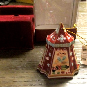 May include: A decorative Christmas ornament shaped like a circus tent. The ornament is red with white snowflake and star designs. It features a painted image of a clown. A red velvet box is in the background.