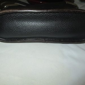 Puede incluir: Un bolso negro con un acabado de grano texturizado y un borde de cuero marr&oacute;n oscuro liso a lo largo de la base. El bolso presenta una forma ovalada alargada y redondeada con costuras visibles en los bordes y accesorios met&aacute;licos.
