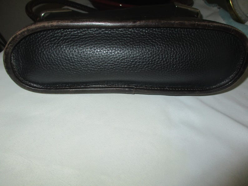 Puede incluir: Un bolso negro con un acabado de grano texturizado y un borde de cuero marr&oacute;n oscuro liso a lo largo de la base. El bolso presenta una forma ovalada alargada y redondeada con costuras visibles en los bordes y accesorios met&aacute;licos.