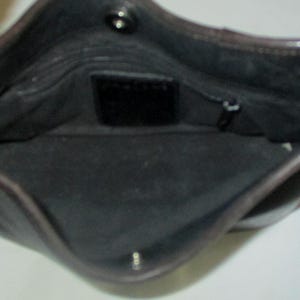 Puede incluir: El interior de un bolso de cuero marr&oacute;n oscuro con forro de tela negra, un cierre de bot&oacute;n a presi&oacute;n en la parte superior y un peque&ntilde;o parche de cuero rectangular centrado en la pared trasera con un bolsillo con cremallera a la derecha.