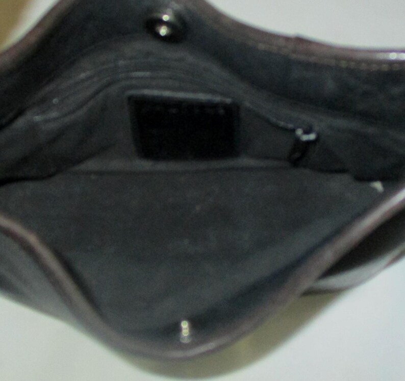 Puede incluir: El interior de un bolso de cuero marr&oacute;n oscuro con forro de tela negra, un cierre de bot&oacute;n a presi&oacute;n en la parte superior y un peque&ntilde;o parche de cuero rectangular centrado en la pared trasera con un bolsillo con cremallera a la derecha.
