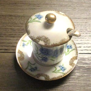 May include: A vintage porcelain sugar bowl with a lid and saucer. The white bowl and lid are decorated with blue floral patterns and gold trim. A small spoon rests on the lid, topped with a gold knob.