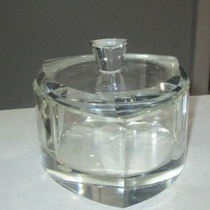 May include: A clear, faceted glass jar with a matching lid. The jar has a heart-shaped base and a triangular knob on the lid. The glass is thick and reflects light, creating a sparkling effect. The jar is likely for decorative or storage purposes.