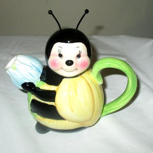 May include: A ceramic teapot shaped like a bee with a black head, yellow body, and green handle. The bee has pink cheeks, black antennae, and holds a light blue flower-shaped spout. The design features painted details on the face and body.