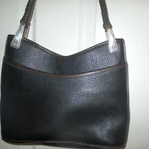 Puede incluir: Un bolso negro texturizado con una correa de hombro trenzada de color marr&oacute;n oscuro. El bolso presenta una abertura superior curva, un bolsillo frontal con ribete marr&oacute;n oscuro y herrajes met&aacute;licos plateados que unen la correa al cuerpo.