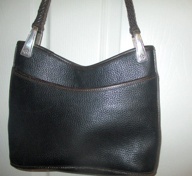 Puede incluir: Un bolso negro texturizado con una correa de hombro trenzada de color marr&oacute;n oscuro. El bolso presenta una abertura superior curva, un bolsillo frontal con ribete marr&oacute;n oscuro y herrajes met&aacute;licos plateados que unen la correa al cuerpo.