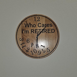May include: A round wooden clock with the text "Who Cares I'm RETIRED" in black. The clock face has black numerals and hands. The clock is hanging on a white wall.
