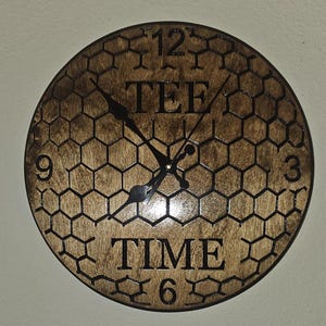 Puede incluir: Reloj de madera redondo con un patrón de panal y las palabras "TEE TIME". Las manecillas del reloj son negras y los números 12, 3, 6 y 9 son visibles. El reloj tiene un acabado marrón oscuro.