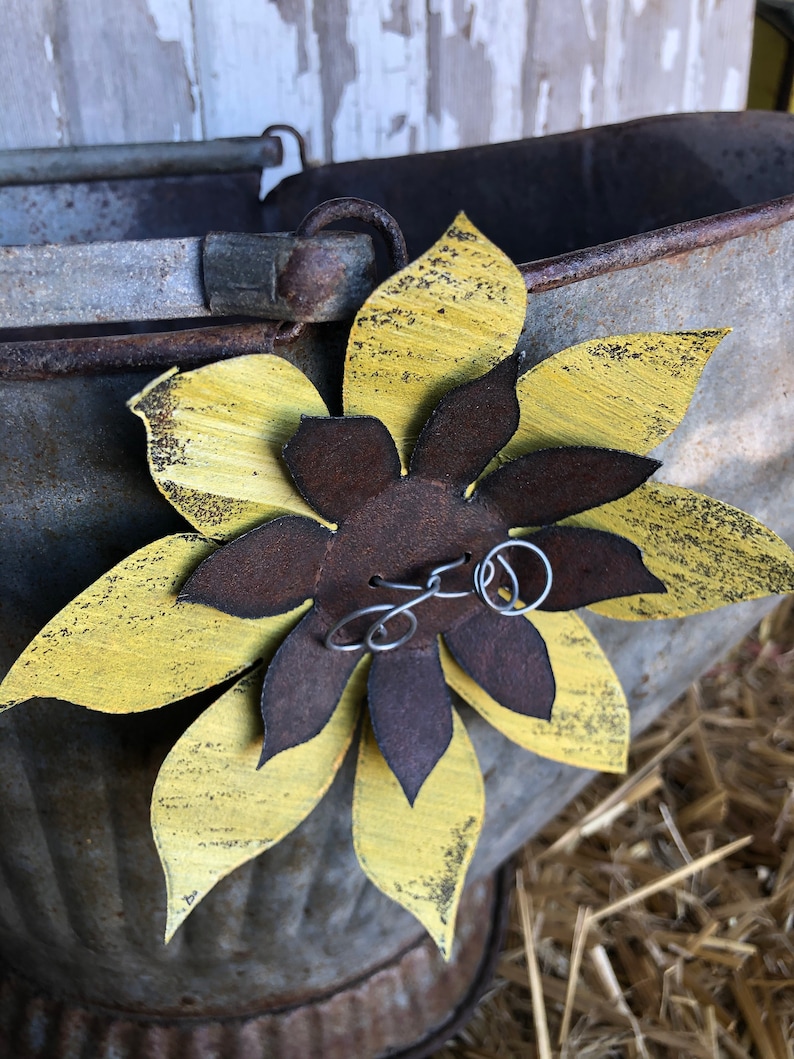 K&ouml;nnte beinhalten: Eine gelbe und braune Metallsonnenblume mit Drahtverzierungen, die an einem verzinkten Metalleimer befestigt ist.