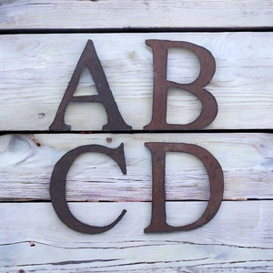 May include: Four metal letters, A, B, C, and D, are arranged in two rows on a light wood background. The letters are rusty brown in color.