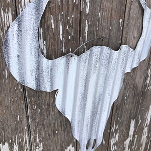 May include: White metal bull skull silhouette with a corrugated texture, hanging on a weathered wooden wall.