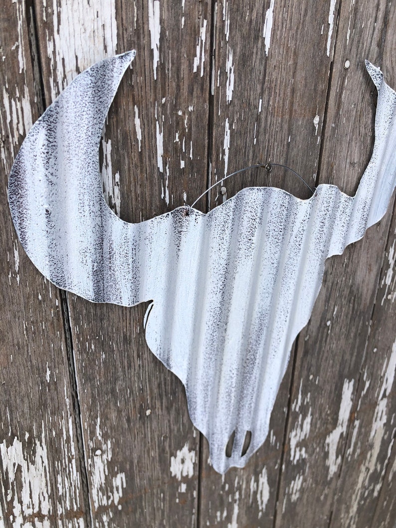 May include: White metal bull skull silhouette with a corrugated texture, hanging on a weathered wooden wall.