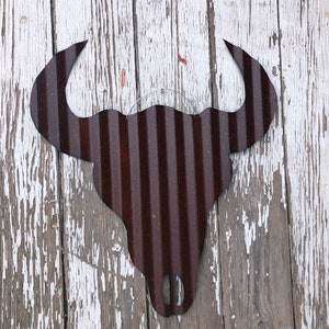 Op de afbeelding: Een rustiek metalen stierenhoofd silhouet met een gegolfde textuur, geschilderd in donkerbruin. Het silhouet is gemonteerd op een verweerd wit houten oppervlak.