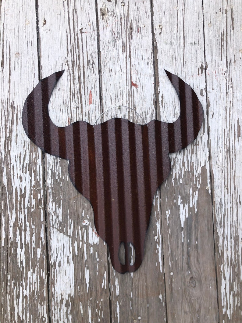 Op de afbeelding: Een rustiek metalen stierenhoofd silhouet met een gegolfde textuur, geschilderd in donkerbruin. Het silhouet is gemonteerd op een verweerd wit houten oppervlak.