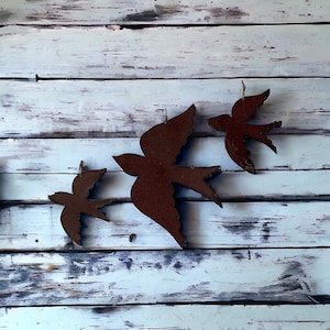 May include: Three rustic metal bird wall decorations in varying sizes. Each bird is a silhouette with a brown, weathered finish, and is suspended by a thin string. The birds are arranged against a backdrop of distressed, white-painted wooden planks.