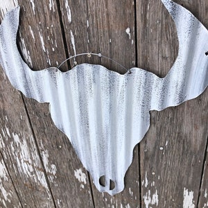 May include: White metal bull skull silhouette with a distressed finish, hanging on a weathered wooden wall.