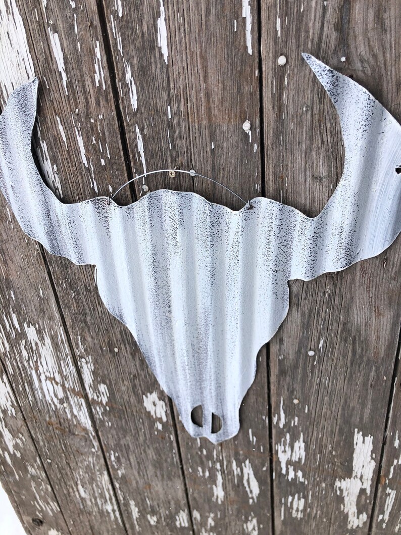 May include: White metal bull skull silhouette with a distressed finish, hanging on a weathered wooden wall.