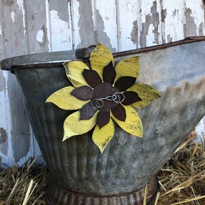 K&ouml;nnte beinhalten: Ein rustikaler Metalleimer mit einer gelben und braunen Sonnenblume, die an der Seite befestigt ist. Der Eimer steht auf einem Strohboden.