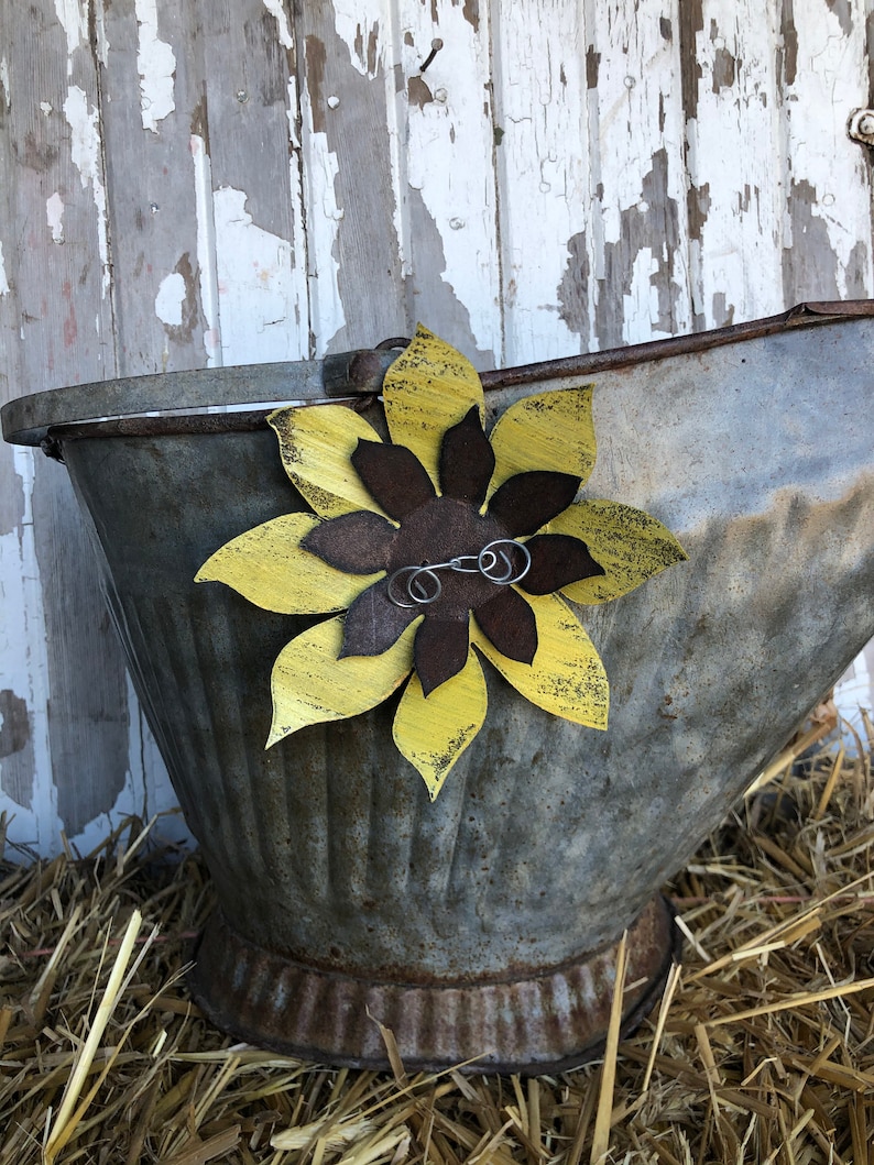 K&ouml;nnte beinhalten: Ein rustikaler Metalleimer mit einer gelben und braunen Sonnenblume, die an der Seite befestigt ist. Der Eimer steht auf einem Strohboden.