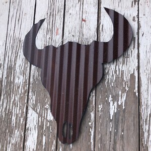 Op de afbeelding: Een bruine metalen stieren schedel silhouet met een gegolfde textuur, hangend aan een witte houten muur.