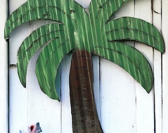 May include: A green and brown wooden palm tree cutout, mounted on a white wooden wall. The palm tree has a rustic, weathered look.