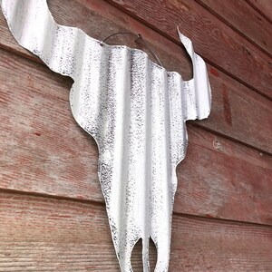 Op de afbeelding: Witte metalen stieren schedel wanddecoratie met een gegolfde textuur. De schedel hangt aan een rode houten muur.