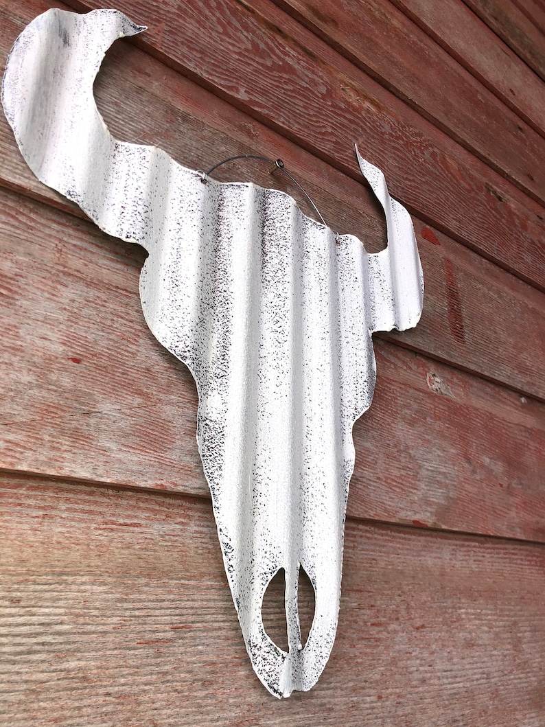 Op de afbeelding: Witte metalen stieren schedel wanddecoratie met een gegolfde textuur. De schedel hangt aan een rode houten muur.