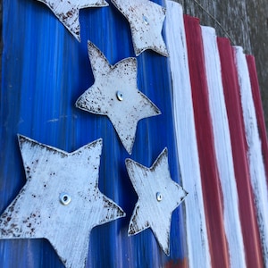 May include: A rustic American flag made of corrugated metal with white stars and red and white stripes. The stars are painted white and have a distressed look. The flag is mounted on a wooden surface.