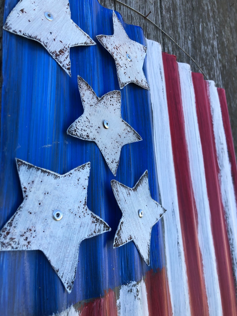 May include: A rustic American flag made of corrugated metal with white stars and red and white stripes. The stars are painted white and have a distressed look. The flag is mounted on a wooden surface.