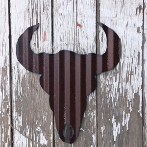 May include: A rustic brown metal bull skull silhouette with a distressed finish. The skull is hanging on a weathered white wooden wall.